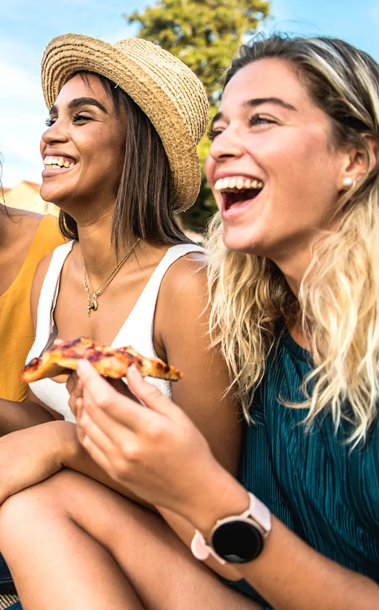 two young women smiling while eating pizza outdoors